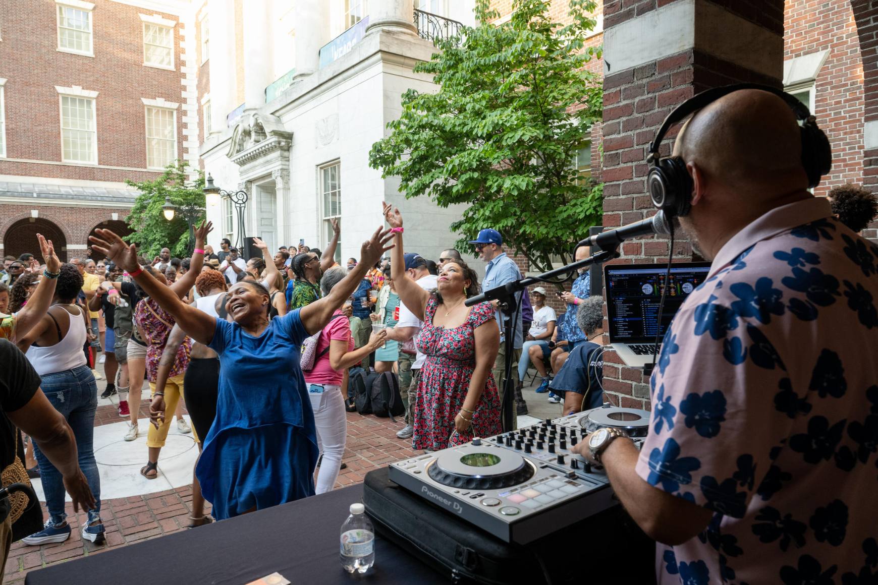 DJ Ted Smooth is at his turntables spinning in the foreground, facing away from the camera. Dancers in front of him throw their arms in the air.